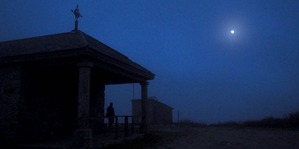 Se vislumbra a la luz de la luna la silueta de la capilla del alto de San Mamede y junto a ella una persona que mira al cielo nocturno